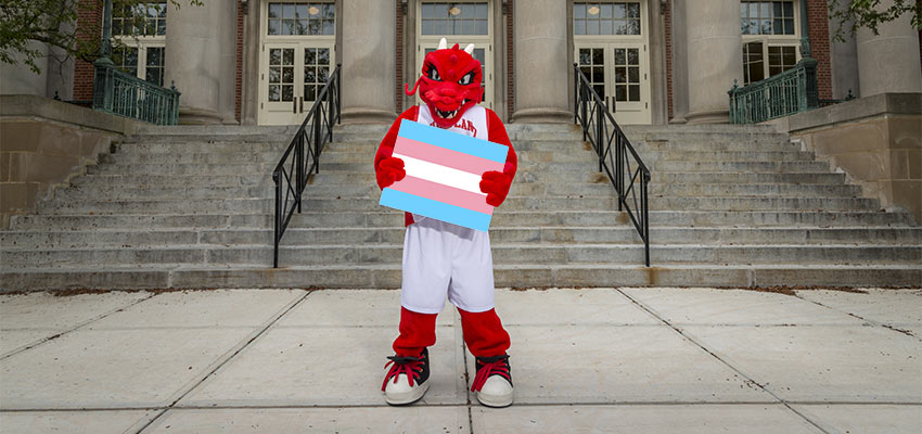 Blaze stands in front of Old Main holding a blue, pink, and white trans flag.