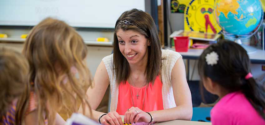 Graduate student sits iwth chidlren at a table in class with a globe and a clock in the background.